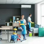 A person wearing a blue uniform and yellow gloves is cleaning a white office desk. They are holding a spray bottle and wiping the desk surface with a cloth, with a computer monitor, phone, and desk lamp visible in the background.