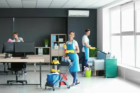 A person wearing a blue uniform and yellow gloves is cleaning a white office desk. They are holding a spray bottle and wiping the desk surface with a cloth, with a computer monitor, phone, and desk lamp visible in the background.