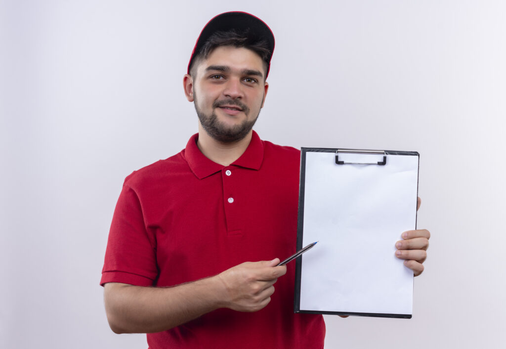 young delivery man in red uniform and cap holding clipboard with blank pages pointing with pen to itasking for signature standing over white background