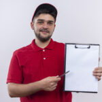 young delivery man in red uniform and cap holding clipboard with blank pages pointing with pen to itasking for signature standing over white background