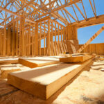 Interior view of a residential timber construction site showing the wooden skeletal frame, roof trusses, and OSB subflooring under a bright blue sky.