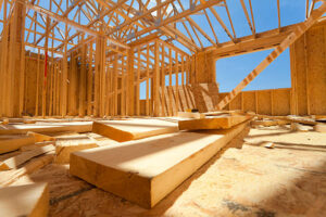 Interior view of a residential timber construction site showing the wooden skeletal frame, roof trusses, and OSB subflooring under a bright blue sky.